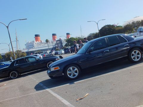 2003 Ford Crown Victoria - dark sedan with chrome wheels sits angled in a striped lot, the historic liner's funnels framed beyond.