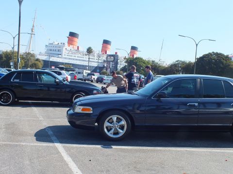 2003 Ford Crown Victoria - Group of men stand between dark full-size sedans, the three-funnel ship dominating the skyline behind.