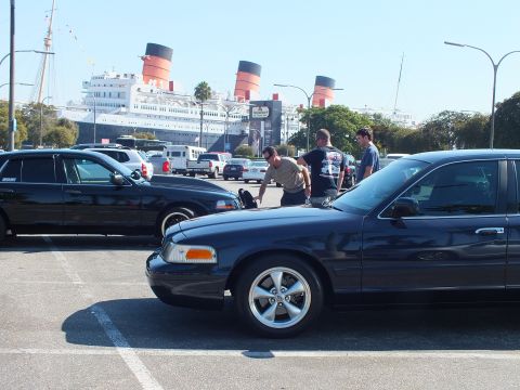 2003 Ford Crown Victoria - Front of a dark sedan with five-spoke wheels, men inspecting another car and the liner across the lot.