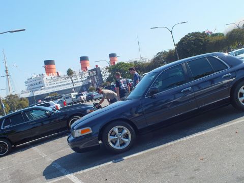 2003 Ford Crown Victoria - Two dark sedans nose to tail in a lot, onlookers and the orange-and-black ship funnels in the distance.
