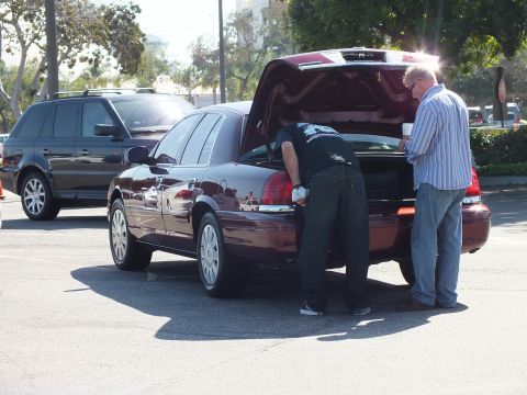 2003 Ford Crown Victoria - One man leans into the open trunk of a maroon sedan while another stands beside it in the bright lot.