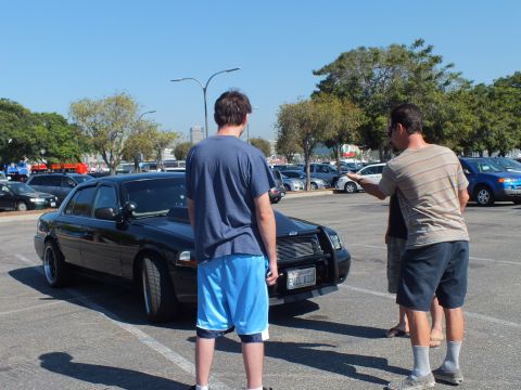2003 Ford Crown Victoria - Men stand looking at a lowered black sedan with chrome grille and a California license plate in the lot.