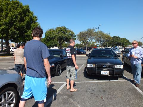 2003 Ford Crown Victoria - Black Ford sedan viewed head-on with California plate, several men standing around cars in the open lot.