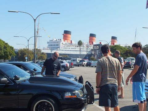 2003 Ford Crown Victoria - man reaches into a black sedan's open door as others talk nearby, the liner's funnels rising behind.