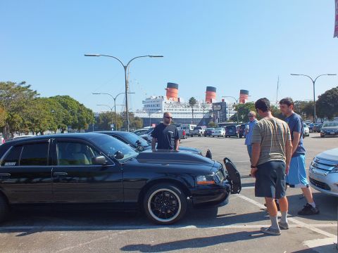2003 Ford Crown Victoria - Black sedan with amber corner lights parked at angle, men in shorts chatting beside the lot and liner.