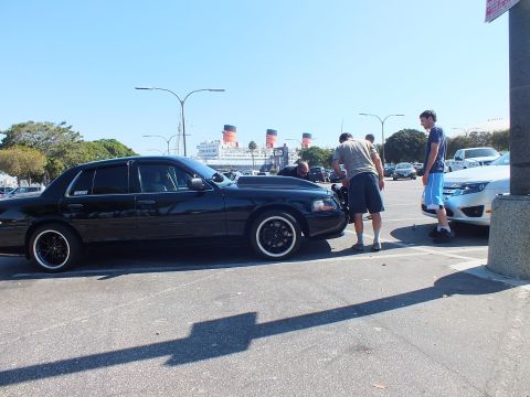 2003 Ford Crown Victoria - Black four-door on raised-white-letter tires, men gathered at the front, ship funnels across the lot.