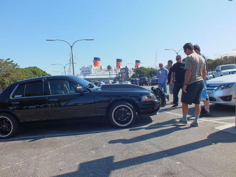 2003 Ford Crown Victoria - Black sedan in profile with black wheels, a cluster of men talking beside it and the liner behind.