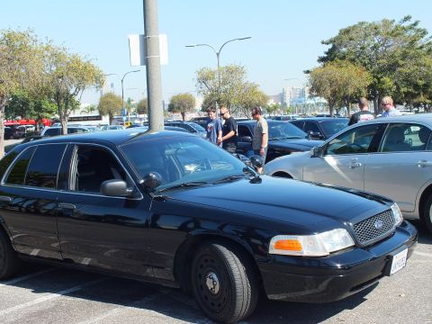 2003 Ford Crown Victoria - Black Ford sedan with chrome grille and amber lights, people and rows of cars filling the lot behind.