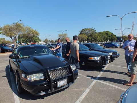 2003 Ford Crown Victoria - Lowered black sedan with steel push bar and scooped hood, plate reading EVIL VIC, men talking beside it.