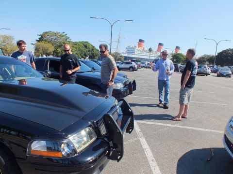2003 Ford Crown Victoria - Men in sunglasses talk over black push-bar sedans, the three-funnel liner moored across the lot.