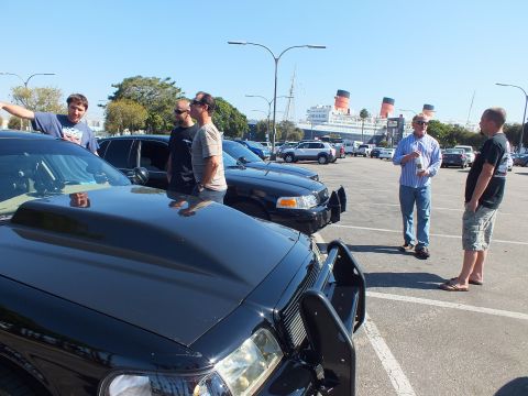 2003 Ford Crown Victoria - black sedan's push bar and hood fill the foreground as men converse, ship funnels rising beyond.