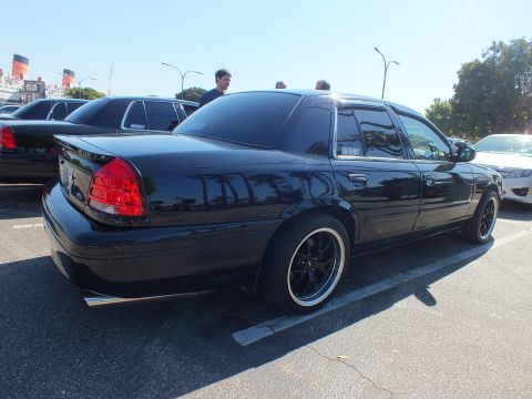 2003 Ford Crown Victoria - Rear of a black four-door sedan with dark multi-spoke wheels and lip spoiler, parked among other cars.