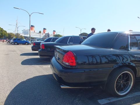 2003 Ford Crown Victoria - Rear three-quarter view of lowered black Crown Victoria sedans parked beside the red-funneled Queen Mary.