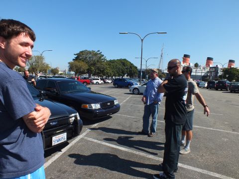 2003 Ford Crown Victoria - group chats in a sunny lot next to a black Crown Victoria, ship funnels in the distance.