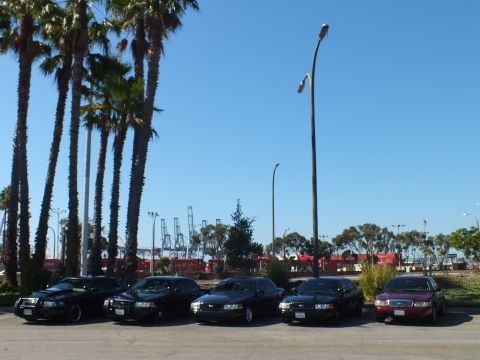 2003 Ford Crown Victoria - Group of full-size sedans in a sunny lot with curved light poles and dockside cranes behind them.