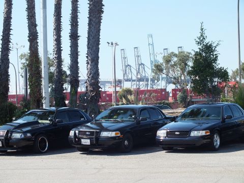 2003 Ford Crown Victoria - Trio of black Crown Victoria sedans, one lowered, parked near red shipping containers and port gantry cranes.