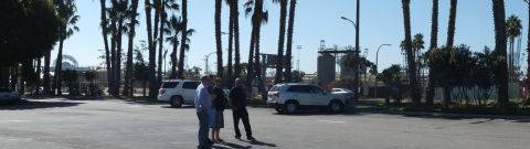 2003 Ford Crown Victoria - Three people talk in a wide paved lot, white SUVs and palm trees with harbor structures behind.
