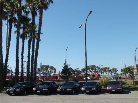 2003 Ford Crown Victoria - Five full-size sedans angled in a lot, tall light pole overhead and red containers across the rail yard.