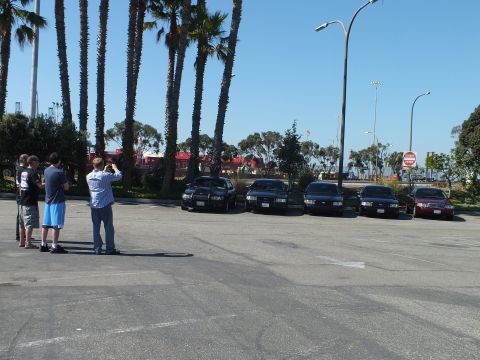 2003 Ford Crown Victoria - Onlookers shoot photos of five lined-up sedans across an open lot, stop sign and palms in view.