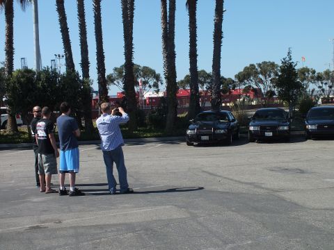 2003 Ford Crown Victoria - Four people stand together photographing parked sedans, palms and red containers framing the scene.