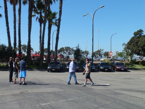 2003 Ford Crown Victoria - Several people cross a paved lot toward parked sedans, one carrying a camera, palms and stop sign behind.