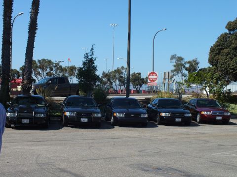 2003 Ford Crown Victoria - Sedan lineup front-on in a lot, a pickup passes behind, Do Not Enter sign and harbor in view.
