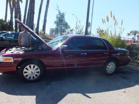 2003 Ford Crown Victoria - Side profile of a dark red Ford Crown Victoria with hood raised in a palm-lined parking lot.