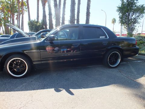 2003 Ford Crown Victoria - Lowered black sedan shown in profile on aftermarket wheels, palms and sky in background.