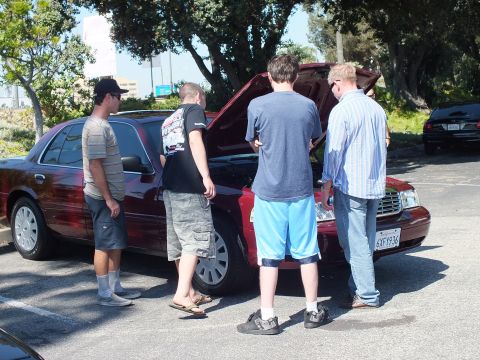 2003 Ford Crown Victoria - Group gathered around a dark red full-size sedan with its hood raised in a parking lot.