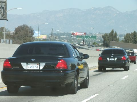 2003 Ford Crown Victoria - pair of black Crown Victorias travel a multi-lane highway toward distant peaks