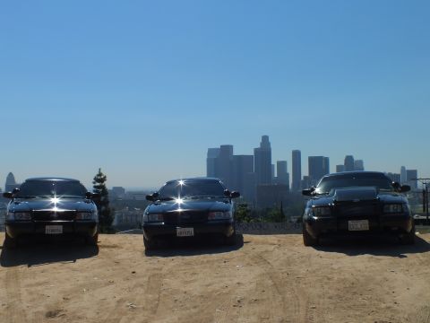 2003 Ford Crown Victoria - Black sedans grouped on a dirt hilltop with downtown towers in the haze