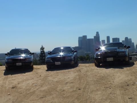 2003 Ford Crown Victoria - Trio of black sedans with headlight glare on a dirt lot facing the city