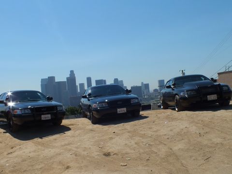 2003 Ford Crown Victoria - Trio of black sedans staged on bare dirt before the city skyline