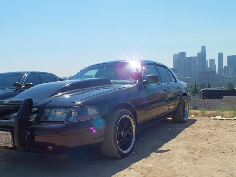 2003 Ford Crown Victoria - Lowered black Ford sedan catches sun flare, black aftermarket wheels and grille guard, second car at left