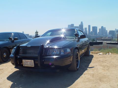 2003 Ford Crown Victoria - Two black Ford sedans on a hilltop dirt lot, the nearer one wearing a push bar and EVIL VTC plate, skyline beyond