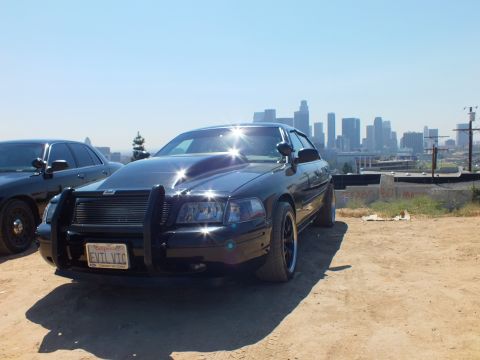 2003 Ford Crown Victoria - Front three-quarter of a blacked-out Ford sedan with grille guard, EVIL VTC plate, city skyline in the distance