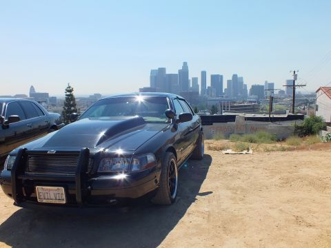 2003 Ford Crown Victoria - Lowered black Ford sedan with push bar parked on a dirt overlook, downtown towers and power lines behind