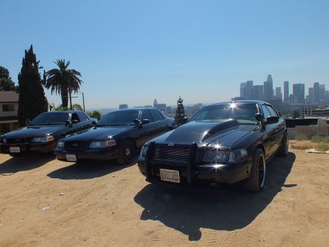2003 Ford Crown Victoria - Trio of blacked-out Crown Victorias staged on a hilltop, palms and downtown skyline behind, push bar on front car