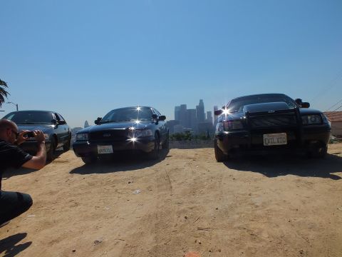 2003 Ford Crown Victoria - Photographer kneels to shoot two black Ford sedans on a dirt lot, downtown skyline framed between them