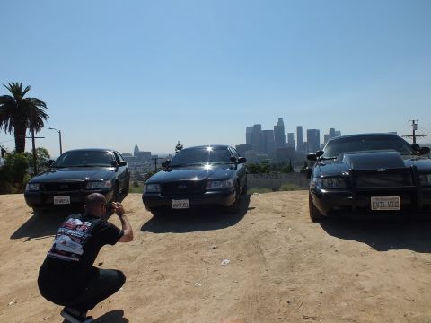 2003 Ford Crown Victoria - man crouches with a camera before three blacked-out Ford sedans, palm and downtown skyline in the haze
