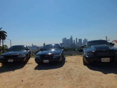 2003 Ford Crown Victoria - Front-on row of three blacked-out Ford sedans, the right one with a push bar and EVIL VTC plate, skyline beyond