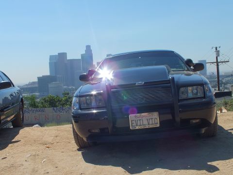 2003 Ford Crown Victoria - Head-on view of a black Ford sedan with push bar and EVIL VTC plate, graffiti wall and downtown towers behind