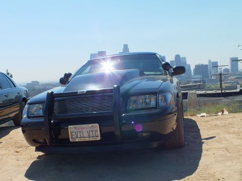 2003 Ford Crown Victoria - Lowered black Ford sedan with grille guard and EVIL VTC plate on a dirt overlook, freeway and skyline behind