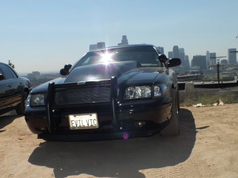 2003 Ford Crown Victoria - Blacked-out Ford sedan with grille guard catches sun flare on the hood, EVIL VTC plate, downtown towers in haze