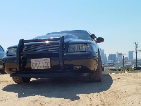 2003 Ford Crown Victoria - Low angle on a blacked-out Ford sedan with push bar and EVIL VTC plate, downtown buildings in the distance