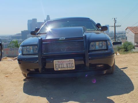 2003 Ford Crown Victoria - Head-on view of a customized black Ford sedan with hood vent, grille guard and EVIL VTC plate, city behind