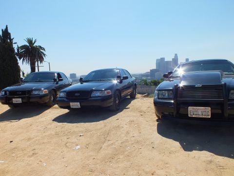 2003 Ford Crown Victoria - Three blacked-out Ford sedans staged on a hilltop, the right one with push bar and EVIL VTC plate, skyline beyond