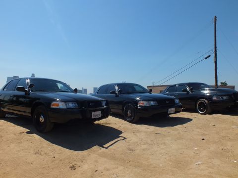 2003 Ford Crown Victoria - Row of three black Ford sedans on a dirt overlook, utility pole at right and faint downtown skyline behind