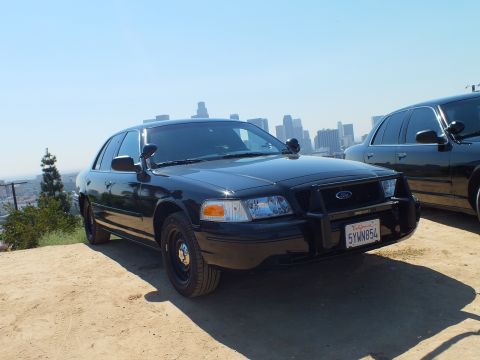 2003 Ford Crown Victoria - Black Ford sedan with grille guard and plate 5YWN854 parked beside a second black car, skyline behind