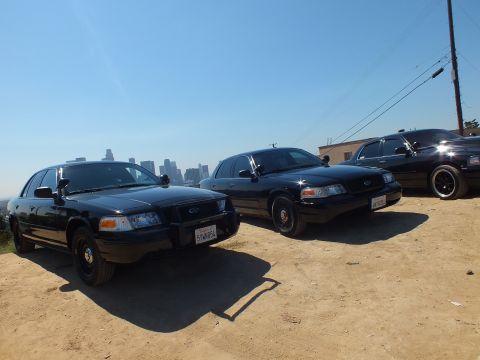 2003 Ford Crown Victoria - Three blacked-out Ford sedans angled on a hilltop, lead plate 5YWN854, downtown skyline and wires behind
